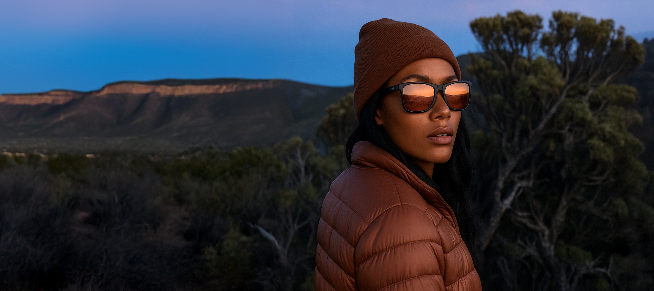 Person wearing a brown puffer jacket and sunglasses in a desert landscape at dusk.